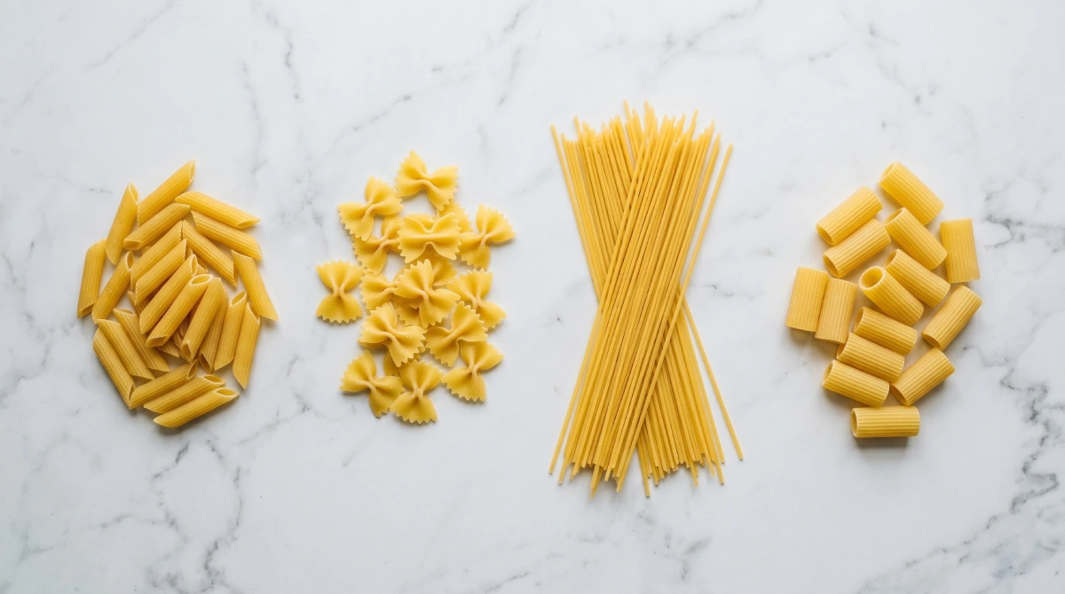 Assorted Italian pasta shapes including penne, farfalle, spaghetti and rigatoni on white marble counter